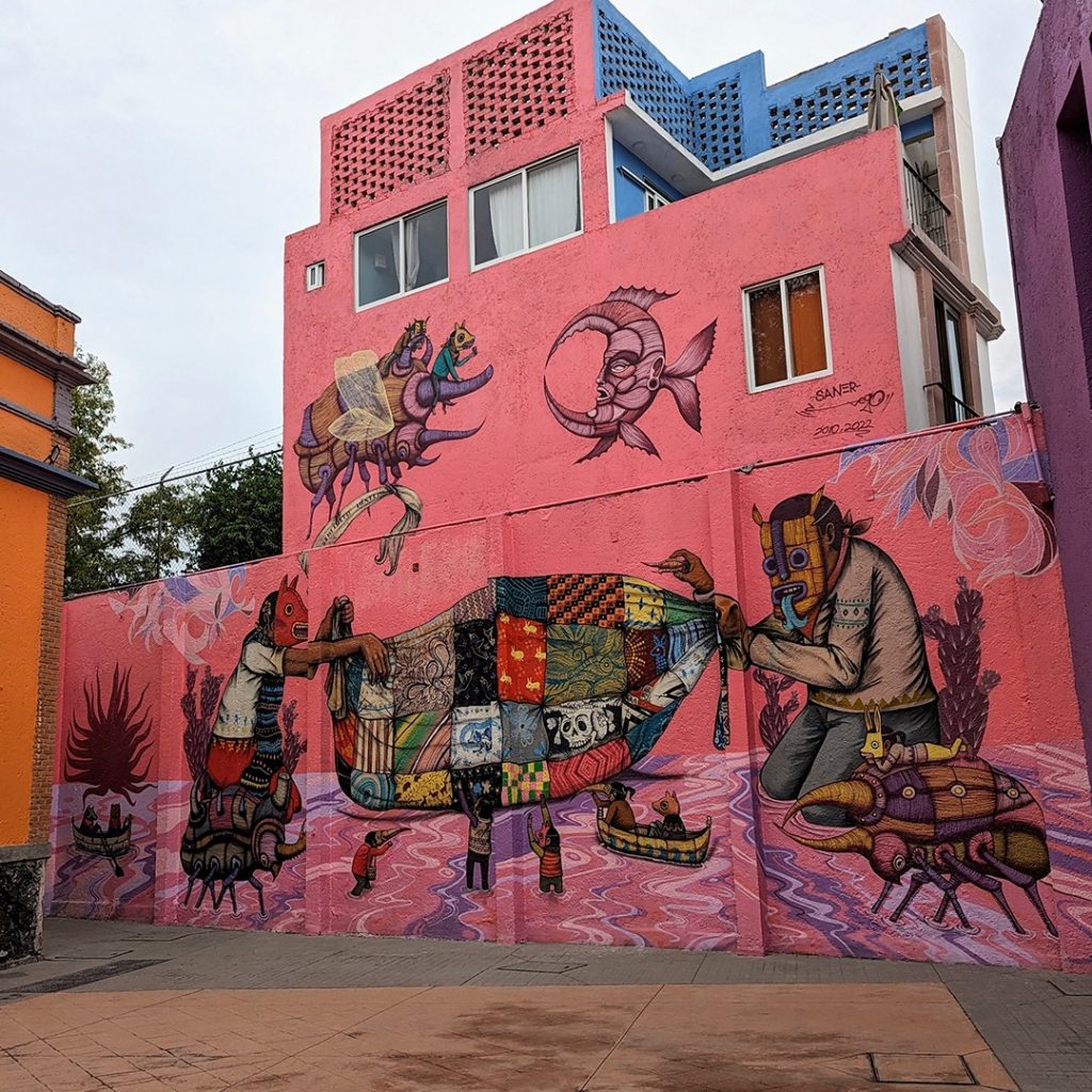 Foto completa del mural “tejedores de sueños”. Tiene una dimensión total de 60 metros cuadrados, está dividido en dos rectángulos, uno más pequeño en la parte superior y otro más grande en el plano inferior y principal. La escena retrata a dos personajes enmascarados, la de la izquierda es una mujer morena arrodillada y que porta una máscara roja que figura a un diablo burlón. La figura de la derecha es un hombre arrodillado que lleva puesta una máscara amarilla. Ambos personajes sostienen una enorme tela que se extiende en el centro del mural y ocupa la mayor parte de nuestra atención. Compuesta por 32 retazos, la tela es elemento central del mural y parece aludir a la diversidad cultural de cada uno de los estados de la República. En la parte superior izquierda observamos a un escarabajo sobrevolar la escena mientras sostiene una manta en sus patas que dice “Infinito México”. Sobre el escarabajo montan dos pequeños niños que portan sendas máscaras amarillas con aspecto animalesco. También en la parte superior, pero del lado derecho encontramos una luna rosácea en cuarto menguante fundida con la figura de un pez. La textura de la misma semeja la de los músculos a carne viva, muestra un rostro antropomorfo con una expresión que denota poder y superioridad.