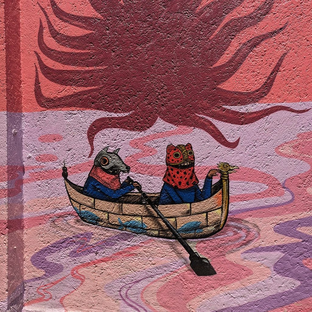 Fotografía de un detalle del Mural. Se trata de dos personajes de menor tamaño, asemejan a niños y portan máscaras de animales con influencia prehispánica. Los dos están dentro de una balsa que navega sobre un cuerpo de agua. Ese detalle del mural se encuentra en la parte inferior, debajo de los dos personajes principales que sostienen la gran tela. A los pies del mural hay una especie de costa con agua en la que varios personajes navegan, algunos semisumergidos en el agua, otros en balsas.