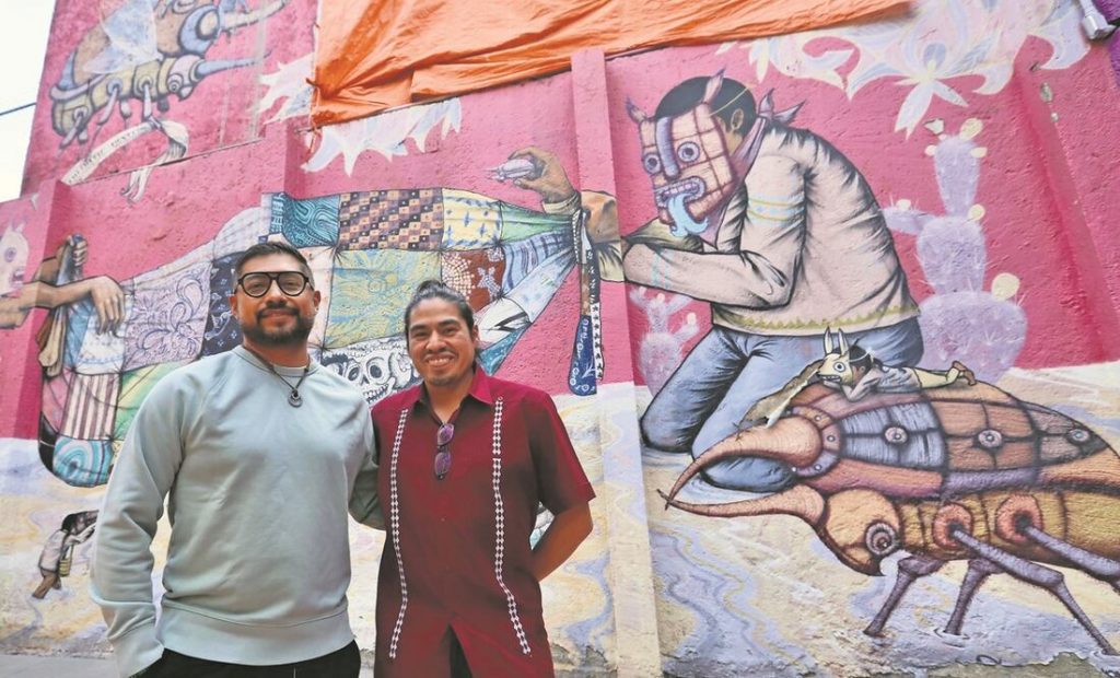 Fotografía de los creadores Edgar “Saner” Flores y Carlos “Sego” Segovia Alanis posando frente al mural “Tejedores de Sueños”.